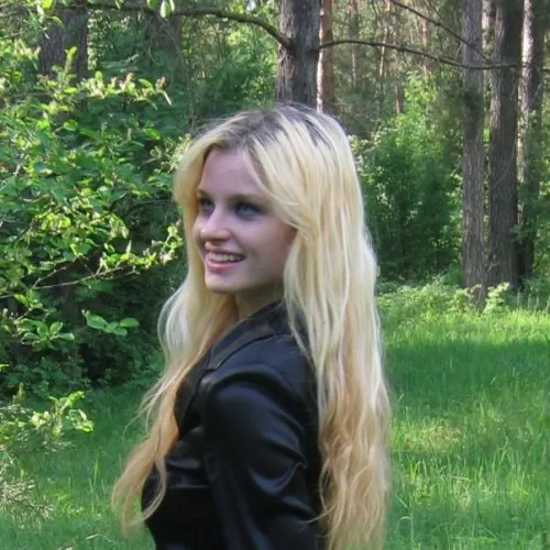 Jeune femme blonde, souriante, elle porte une veste noire en forêt