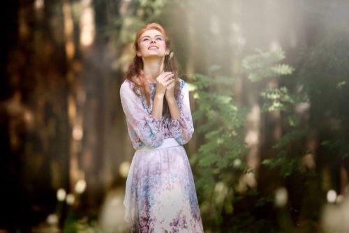 Une femme en robe à fleur se serre les mains en signe de plaisir. Elle a une ronde à fleur dans une forêt