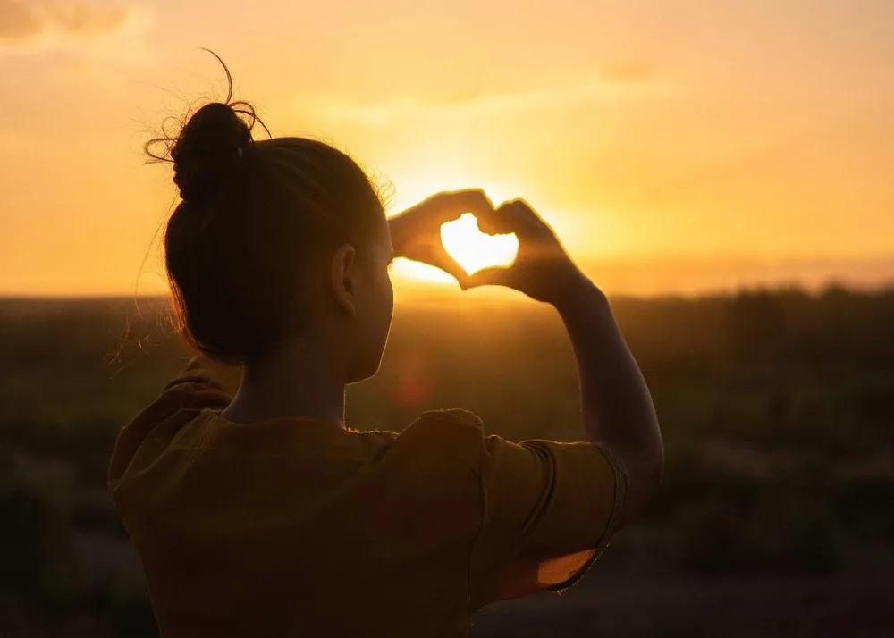 Une jeune femme fait un cœur avec ses doigts et on voit le soleil à travers